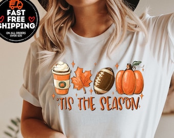 May include: White t-shirt with a graphic design of a coffee cup, a leaf, a football, and a pumpkin. The text "Tis the Season" is written below the graphic.