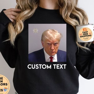May include: Black sweatshirt with a photo of Donald Trump in a mugshot. The text "CUSTOM TEXT" is printed below the photo.