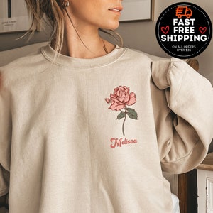 May include: Beige sweatshirt featuring a pink rose graphic with green leaves and the name "Melissa" in a cursive font. The sweatshirt has long sleeves and a crew neck. Additional text includes "MORE THAN 10 COLORS + SIZES".