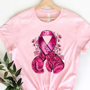 May include: Pink t-shirt with a pink ribbon and boxing gloves graphic. The ribbon has a leopard print pattern and is surrounded by pink flowers.