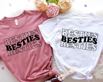 May include: Two t-shirts with the word "Besties" printed on them in a retro style font. The shirts are white and pink.
