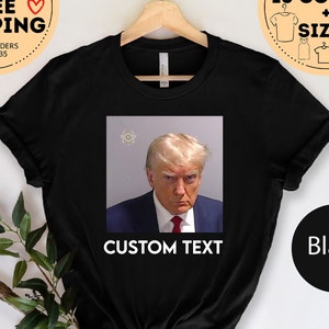 May include: Black t-shirt with a photo of Donald Trump in a mugshot. The text "CUSTOM TEXT" is printed below the photo.