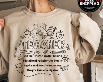 May include: Beige sweatshirt with a black graphic design that says "TEACHER" and includes a definition of the word. The definition includes the words "multi-tasking", "educational rockstar", "inspire", and "encourage".