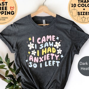 May include: Dark gray heather t-shirt with the text "I CAME I SAW I HAD ANXIETY SO I LEFT" in pastel colors and floral accents. The shirt is displayed on a wooden hanger.