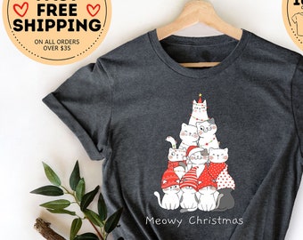 Può includere: T-shirt grigio scuro mélange con un design a forma di albero di Natale fatto di gatti. I gatti indossano cappelli e sciarpe rosse. Il testo "Meowy Christmas" è stampato sotto. La maglietta è esposta su una gruccia di legno.