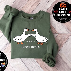Puede incluir: Una sudadera verde con un dise&ntilde;o de ganso blanco y el texto "Goose Bumps".