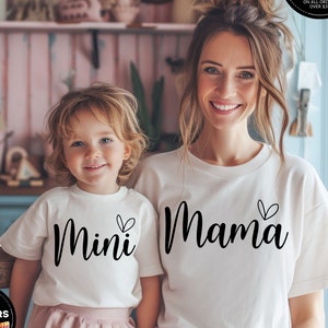 May include: White t-shirts for a parent and child. The adult shirt says "Mama" with a small heart and the child's shirt says "Mini" with a small heart.
