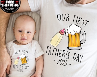 May include: White t-shirt with the text "Our First Father's Day - 2023 -" and a graphic of a baby bottle pouring into a beer mug. A white onesie with the same text and graphic is shown on a baby.