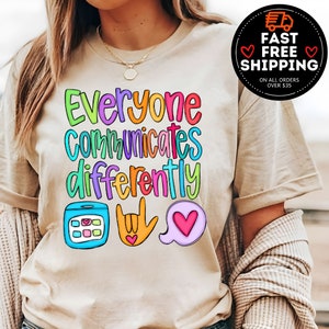 May include: A beige t-shirt with the text "Everyone Communicates Differently" in rainbow colors. The text is surrounded by three icons: a phone with a grid of squares, a hand making the sign for "I love you" in American Sign Language, and a speech bubble with a heart inside.
