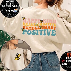 May include: A beige sweatshirt with a rainbow-colored graphic that says "Happy Kind Brave Creative Humble Smart Positive" with bee illustrations. The sweatshirt is shown on a model wearing blue jeans.