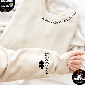 May include: A beige sweatshirt with the words "autism mom" in black script across the chest. The sleeve features the name "William" and a puzzle piece. The image also shows a graphic with text that says "More than 10 colors + sizes."