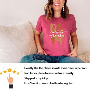 May include: A magenta t-shirt with rolled sleeves. The shirt features the words "represents freedom" in a gold and white script design. The shirt is made of soft fabric.