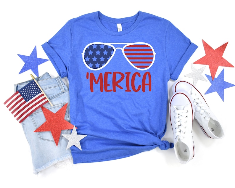 Puede incluir: Una camiseta azul con un gr&aacute;fico rojo, blanco y azul de gafas de sol con el texto 'MERICA' en rojo.