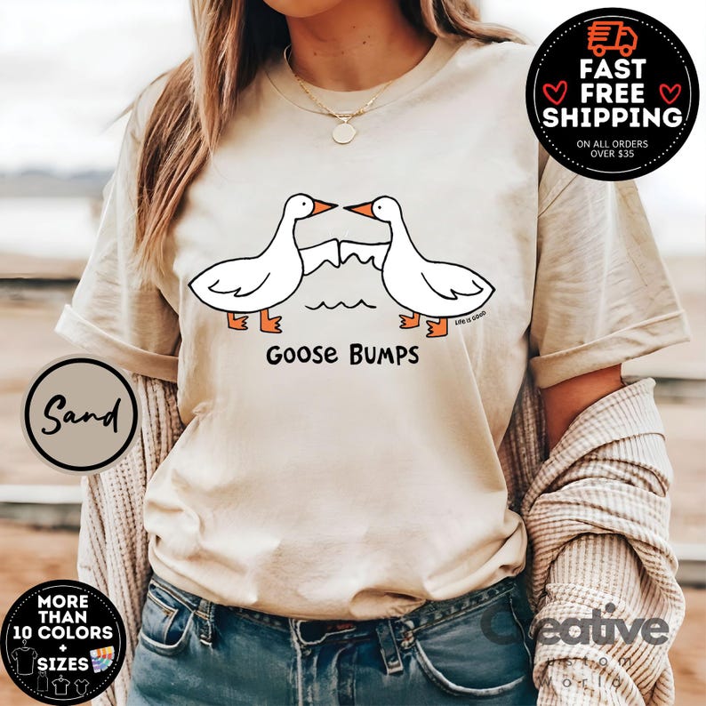 以下が含まれることがあります： ベージュのTシャツに、向かい合った2羽の漫画のガチョウのグラフィックがプリントされています。下に「Goose Bumps」という文字がプリントされています。このTシャツは、青いジーンズを履いた人が着用しています。