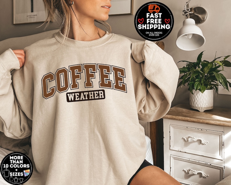 May include: A beige sweatshirt with the text "COFFEE WEATHER" in brown letters. The sweatshirt is a relaxed fit and has a crew neck.