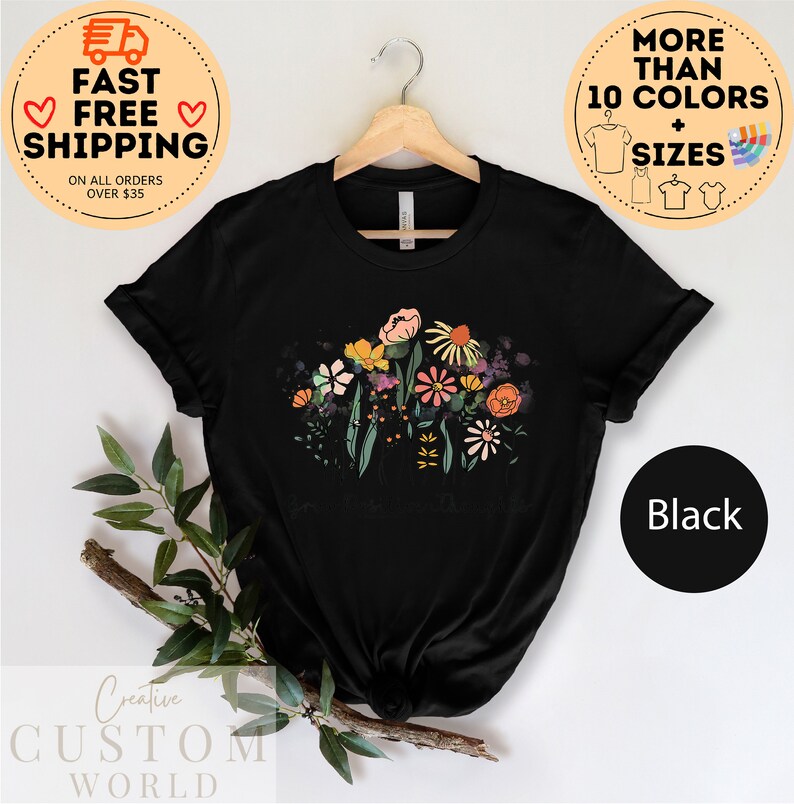 May include: Black t-shirt featuring a floral design with a bouquet of colourful flowers and green leaves. The shirt is on a hanger with a wooden handle.