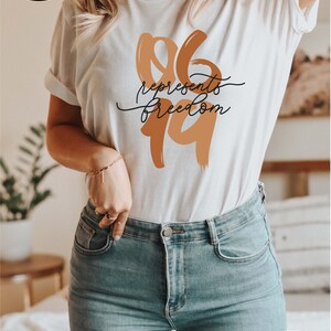 May include: Cream-colored t-shirt with the text "96 represents freedom" in brown and black. The shirt is paired with light blue jeans. The image also includes promotional graphics for fast, free shipping and more color and size options.