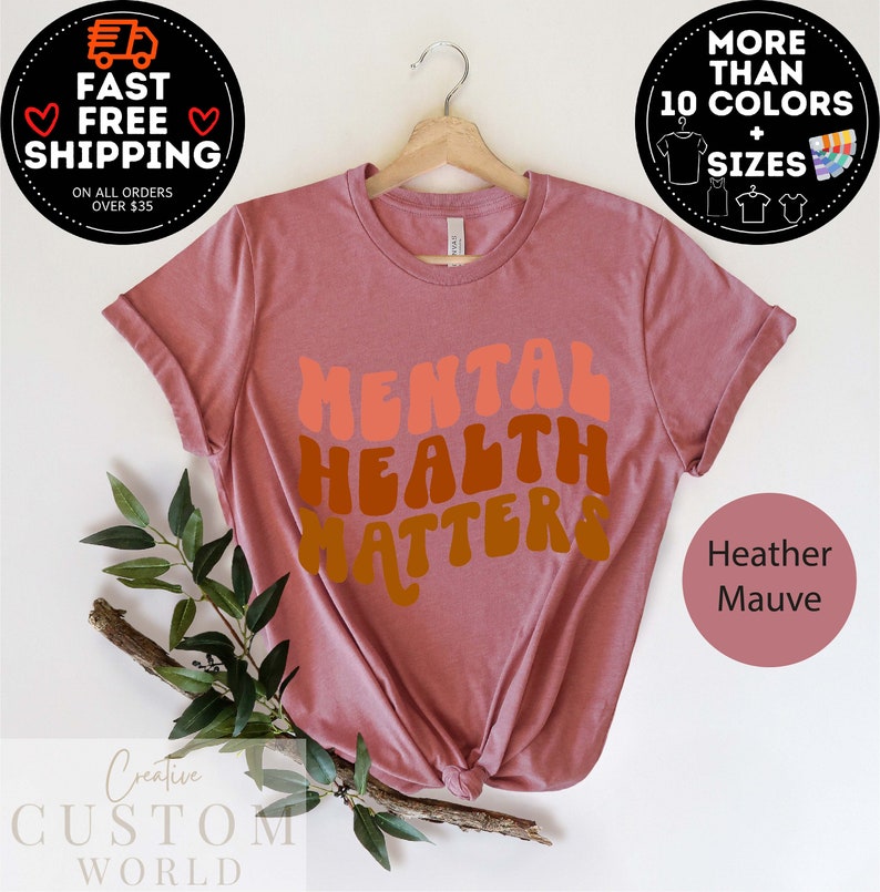 May include: A heather mauve t-shirt with the text "MENTAL HEALTH MATTERS" in a retro font style. The text is in shades of orange, pink, and brown.