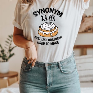 K&ouml;nnte beinhalten: Wei&szlig;es T-Shirt mit einer schwarzen Grafik, auf der "Synonym Rolls" steht, darunter ein Cartoon-Zimtschnecken. Der Text unter der Zimtschnecke lautet "Just like grammar used to make."