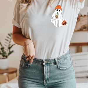 May include: White t-shirt featuring a graphic of a ghost wearing an orange beanie and holding a pumpkin and a cup of coffee. The text "MORE THAN 10 COLORS + SIZES" is in a circle with a rainbow of colours and a variety of t-shirts.