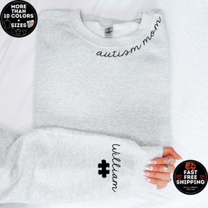 May include: A light gray sweatshirt with the words "autism mom" in black script across the chest. The sleeve features the name "William" and a black puzzle piece. The image also includes text that says "More than 10 colors + sizes".
