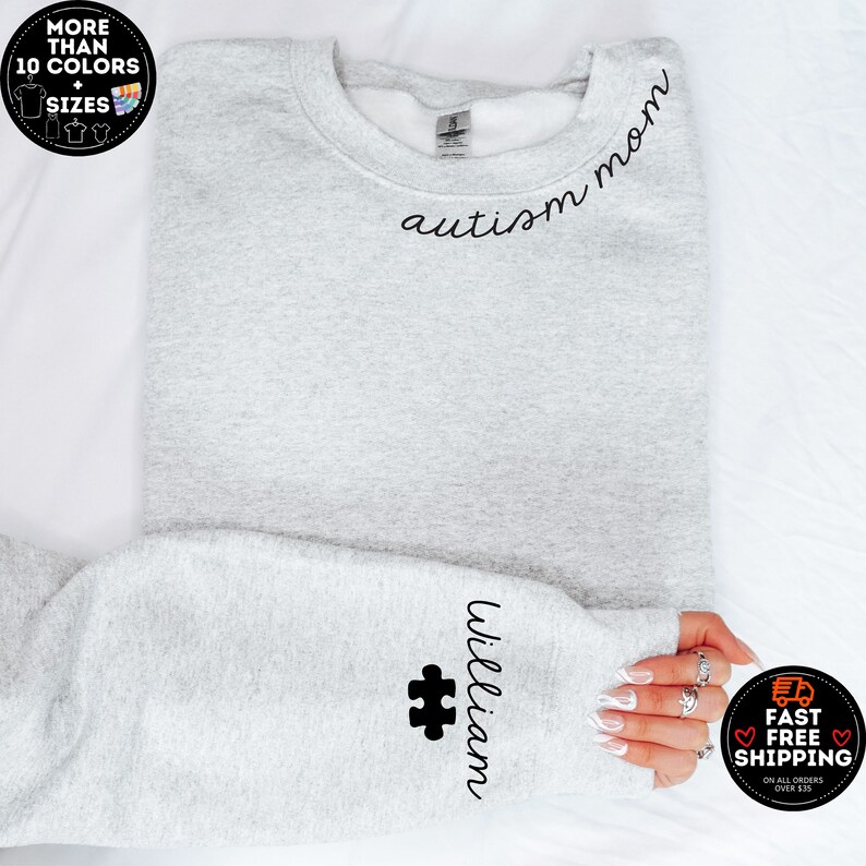 May include: A light gray sweatshirt with the words "autism mom" in black script across the chest. The sleeve features the name "William" and a black puzzle piece. The image also includes text that says "More than 10 colors + sizes".