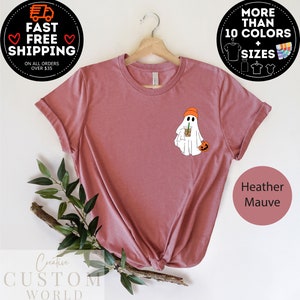 May include: Heather mauve t-shirt featuring a white ghost wearing an orange beanie, holding a pumpkin and a coffee cup. The text "Creative Custom World" is at the bottom of the image.