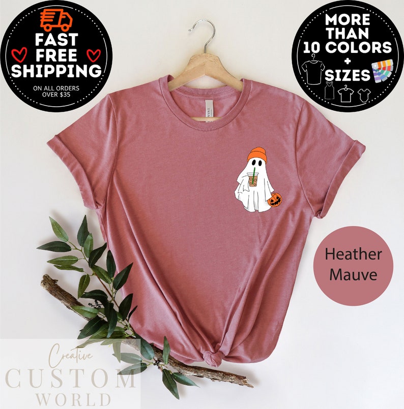 May include: Heather mauve t-shirt featuring a white ghost wearing an orange beanie, holding a pumpkin and a coffee cup. The text "Creative Custom World" is at the bottom of the image.