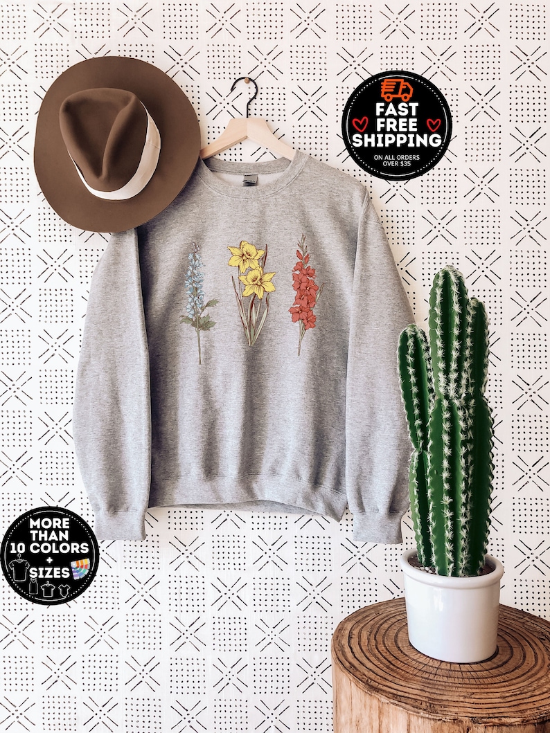 May include: A heather gray sweatshirt featuring embroidered botanical illustrations of blue, yellow, and red flowers. The sweatshirt is displayed on a hanger with a brown hat to the left. The image also includes a cactus in a white pot and text that says "MORE THAN 10 COLORS + SIZES".