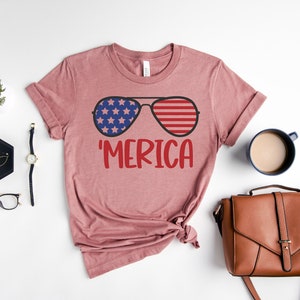 Puede incluir: Una camiseta rosa con un gr&aacute;fico de gafas de sol con la bandera estadounidense en las lentes y la palabra 'MERICA' debajo.