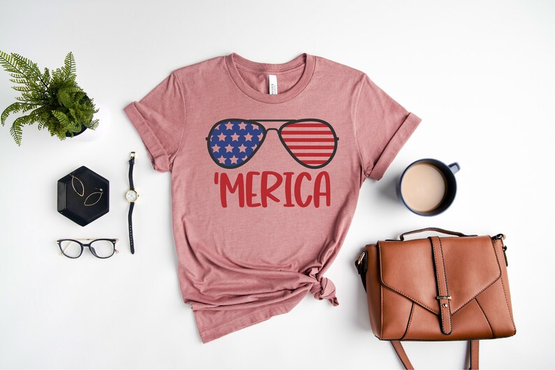 Puede incluir: Una camiseta rosa con un gr&aacute;fico de gafas de sol con la bandera estadounidense en las lentes y la palabra 'MERICA' debajo.