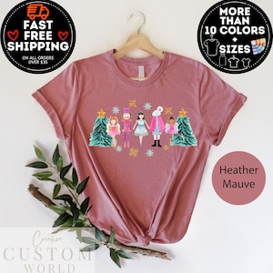 May include: A heather mauve t-shirt with a Christmas-themed design featuring a nutcracker, a mouse, and ballerinas. The design is in a watercolor style and includes a Christmas tree, snowflakes, and stars.