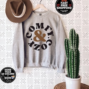 May include: Grey sweatshirt with the text "Comfy & Cozy" in a brown and black circle design. The sweatshirt is hanging on a hanger against a white background with a black and white geometric pattern.