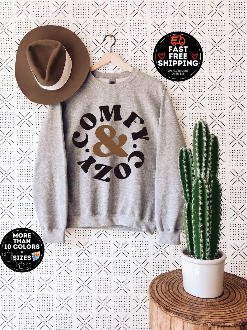 May include: Grey sweatshirt with the text "Comfy & Cozy" in a brown and black circle design. The sweatshirt is hanging on a hanger against a white background with a black and white geometric pattern.