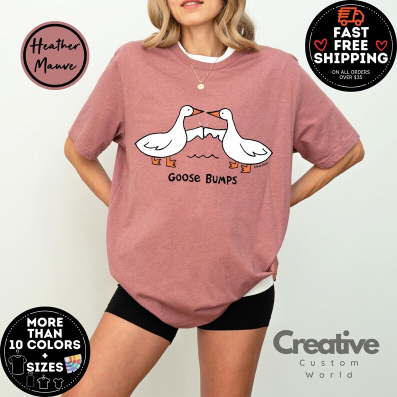 以下が含まれることがあります： ヘザーモーヴのTシャツに、拳をぶつけ合う2羽のガチョウのグラフィックがプリントされています。ガチョウの下には「Goose Bumps」というテキストがプリントされています。