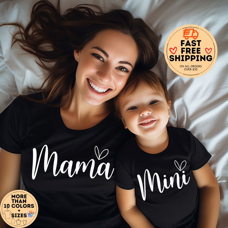 May include: A black t-shirt with the text "Mama" and a heart design, and a black baby onesie with the text "Mini" and a heart design. The shirts are worn by a woman and a baby lying on a white bed.
