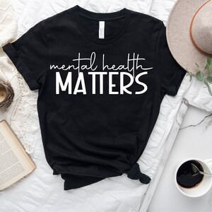 May include: Black t-shirt with white text that reads "mental health matters". The shirt is laid flat on a white surface with a book, a clock, a cup, and a hat.