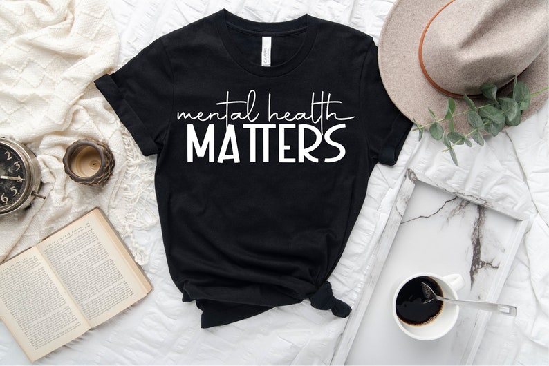 May include: Black t-shirt with white text that reads "mental health matters". The shirt is laid flat on a white surface with a book, a clock, a cup, and a hat.