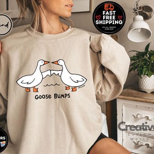 Puede incluir: Una sudadera de color beige claro con un gr&aacute;fico de dos gansos que se miran con el texto "Goose Bumps" impreso debajo.