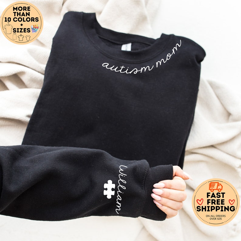 May include: Black sweatshirt with the words "autism mom" in white script across the chest. The sleeve features the name "William" and a puzzle piece graphic. The sweatshirt is folded on a light-colored surface.