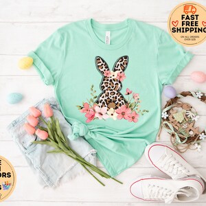 May include: A light green t-shirt with a graphic of a leopard print bunny with pink flowers. The shirt is on a white surface with a pair of white sneakers and a pair of light blue denim shorts.