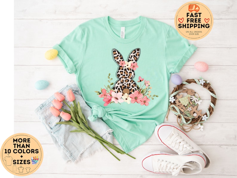 May include: A light green t-shirt with a graphic of a leopard print bunny with pink flowers. The shirt is on a white surface with a pair of white sneakers and a pair of light blue denim shorts.