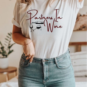Puede incluir: Camiseta blanca con el texto "Partners In Wine" en escritura marr&oacute;n oscuro. Una ilustraci&oacute;n en blanco y negro de una botella de vino vertiendo en una copa est&aacute; por encima del texto.