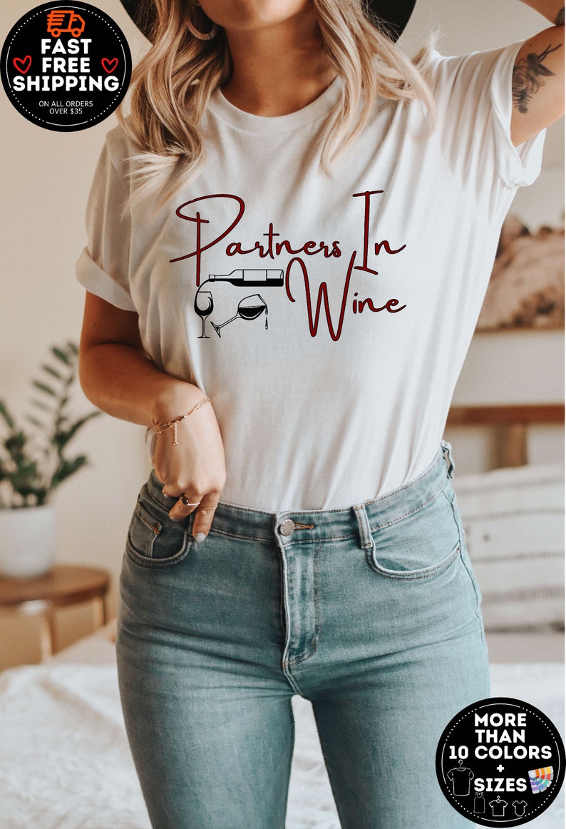 Puede incluir: Camiseta blanca con el texto "Partners In Wine" en escritura marr&oacute;n oscuro. Una ilustraci&oacute;n en blanco y negro de una botella de vino vertiendo en una copa est&aacute; por encima del texto.