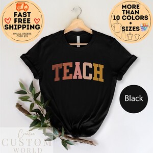 Może przedstawiać: Czarny T-shirt z napisem "TEACH" w kolorze r&oacute;żowym, pomarańczowym i brązowym. Koszulka jest zawieszona na drewnianym wieszaku.