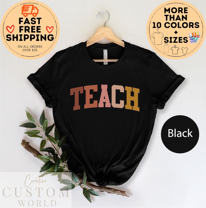 Może przedstawiać: Czarny T-shirt z napisem "TEACH" w kolorze r&oacute;żowym, pomarańczowym i brązowym. Koszulka jest zawieszona na drewnianym wieszaku.