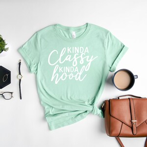 Puede incluir: Una camiseta verde menta con el texto "Kinda Classy Kinda Hood" en escritura blanca. La camiseta est&aacute; combinada con un bolso marr&oacute;n, un reloj, gafas y una taza de caf&eacute; sobre una superficie blanca.
