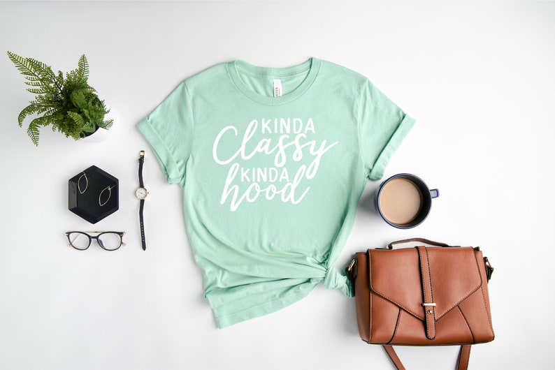 Puede incluir: Una camiseta verde menta con el texto "Kinda Classy Kinda Hood" en escritura blanca. La camiseta est&aacute; combinada con un bolso marr&oacute;n, un reloj, gafas y una taza de caf&eacute; sobre una superficie blanca.