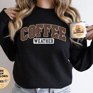 May include: Black sweatshirt with the text "COFFEE WEATHER" in brown letters. The sweatshirt is being modeled by a person holding a white mug with the text "FAST FREE SHIPPING" in orange and red.