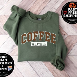 May include: A green sweatshirt with the text "COFFEE WEATHER" in brown letters. The sweatshirt is folded and laid on a wood surface.